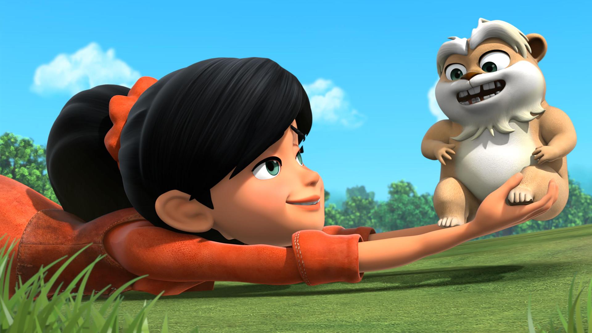 Scarlett liegt auf dem Bauch vor blauem Himmel auf einer grünen Wiese und hält Hamster Derke in ihren nach vorne ausgestreckten Händen. Beide lachen fröhlich.