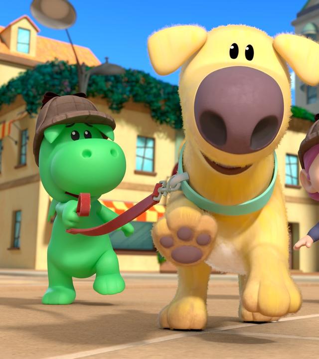 Drache Grisu und seine Freundin Stella jagen mit dem Hund Dinki durch eine Straße von Drachenstadt. Der Hund Dinki rennt voraus. Grisu und Stella tragen Mützen wie Sherlock Holmes.