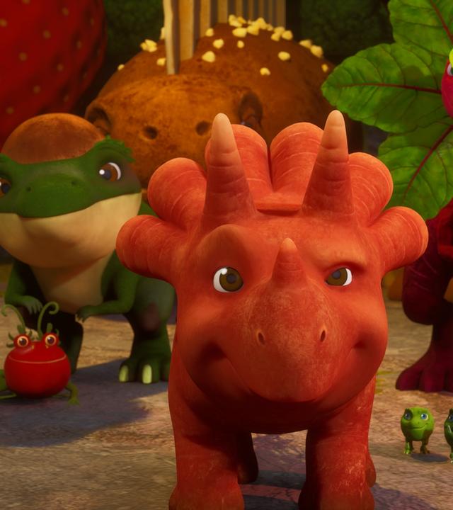 Das Trikarottops-Mädchen Ginger (es sieht aus wie eine orangefarbene Karotte mit Gesicht und Beinen.) steht in der Bildmitte und schaut nach vorne. Um sie herum stehen viele Vegesaurier dicht an dicht: Ein Avocadodon, ein Rotrüben-Saurier, ein Tomatodon, die Erbs-Rexe und noch viele weitere etwas im HIntergrund.