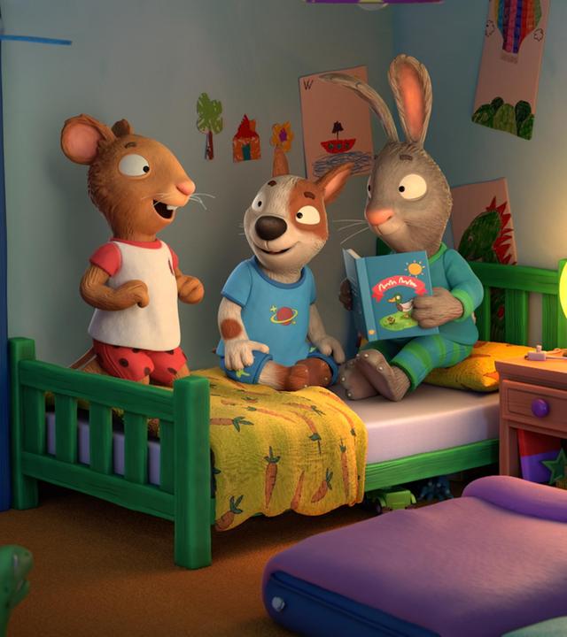 Abends im Zimmer während einer Übernachtungsparty: Die drei Freunde Posy, die Maus, Zac, der Hund und Pip, das Kaninchen haben es sich gemeinsam auf einem Bett gemütlich gemacht. Pip liest eine Geschichte aus einem Buch vor. Zwei Gästebetten aus Luftpolstermatratzen liegen im Zimmer auf dem Boden. Auf dem Nachttisch leuchtet ein Mond als Nachttischlampe.