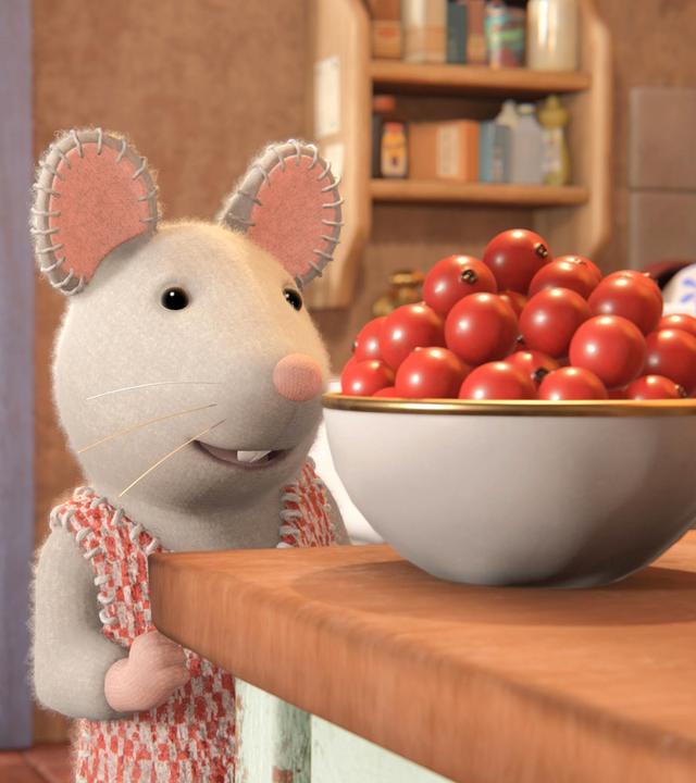 Julia steht in der Küche vor einer Schüssel mit roten Beeren auf einem Tisch und schaut sie begehrlich an. Hinter ihr verlässt Sam gerade die Küche. Er ist von hinten zu sehen und geht auf einen Tisch im Nebenraum zu.