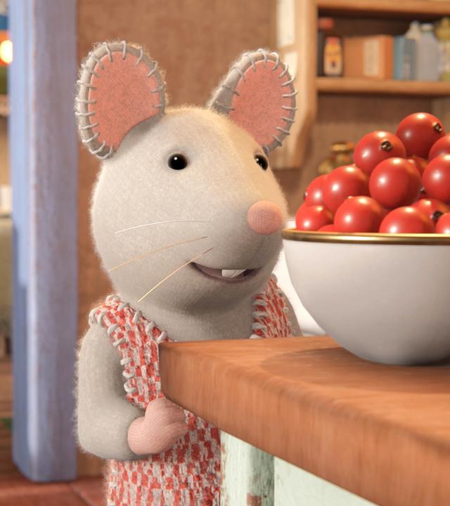 Julia steht in der Küche vor einer Schüssel mit roten Beeren auf einem Tisch und schaut sie begehrlich an. Hinter ihr verlässt Sam gerade die Küche. Er ist von hinten zu sehen und geht auf einen Tisch im Nebenraum zu.