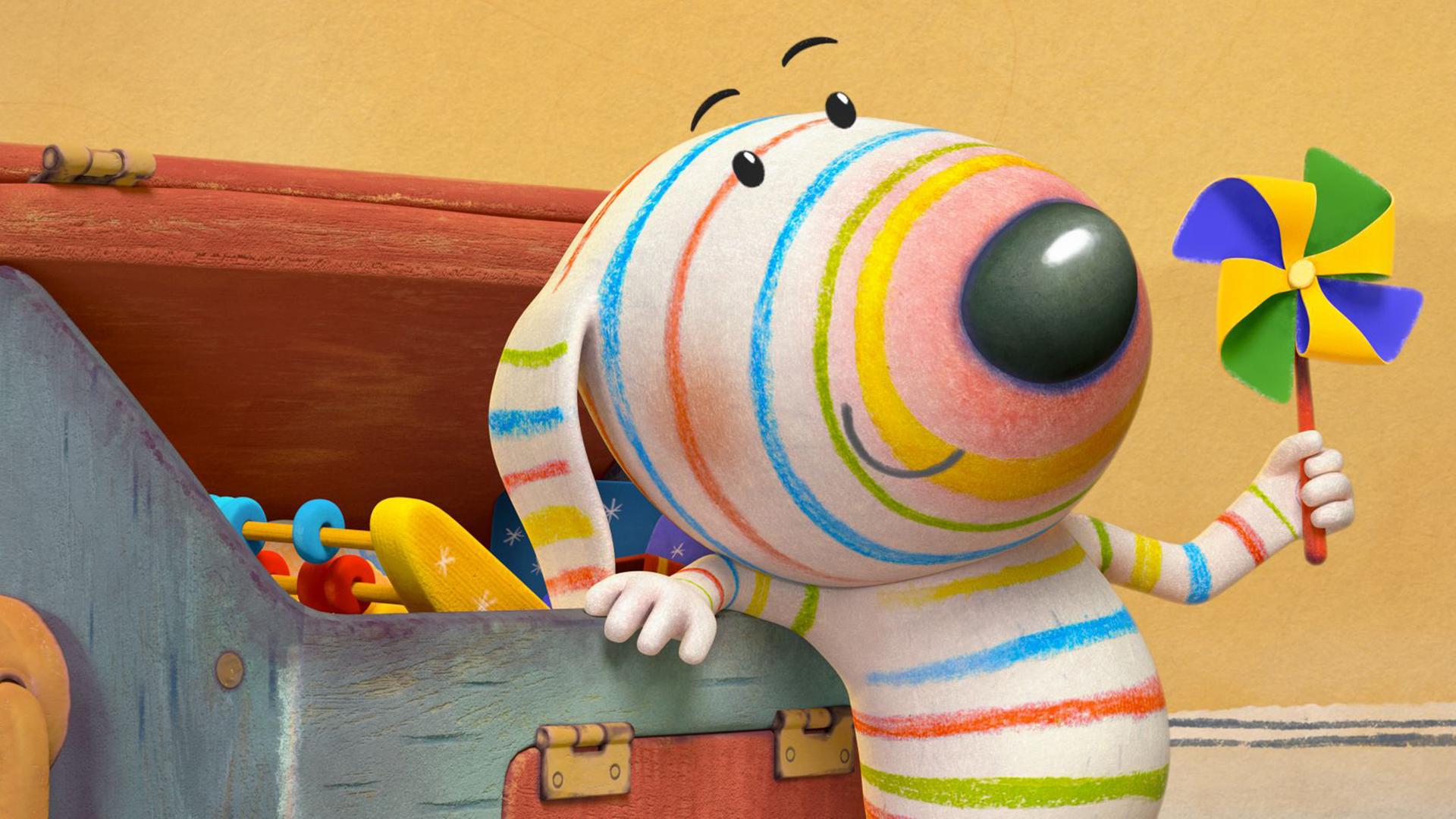 Dylan steht angelehnt an seine geöffnete Spielkiste. In einer Pfote hält er ein Mini-Windrad hoch, das wie eine bunte Blume aussieht. Aus der Kiste ragt ein Rechenschieber hervor.