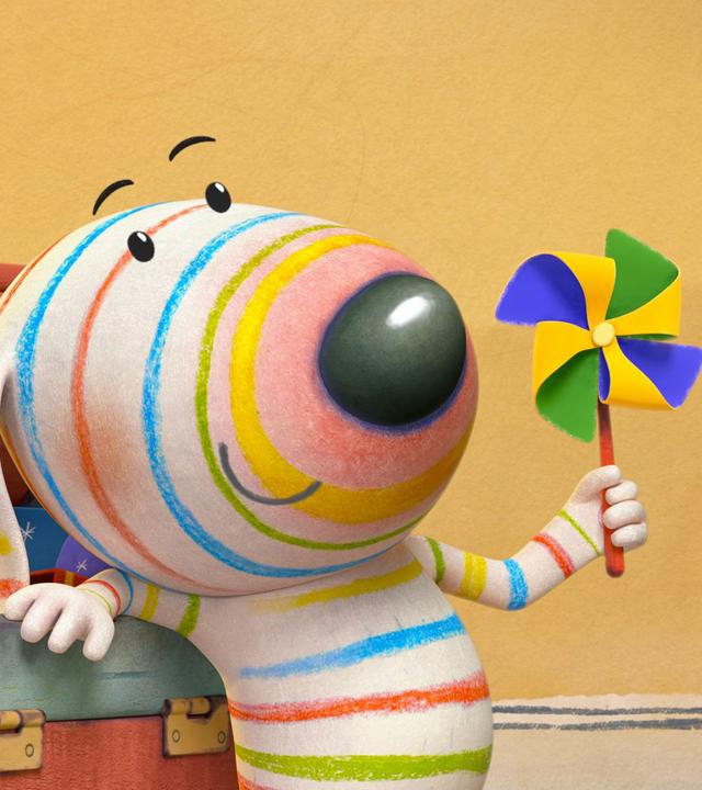 Dylan steht angelehnt an seine geöffnete Spielkiste. In einer Pfote hält er ein Mini-Windrad hoch, das wie eine bunte Blume aussieht. Aus der Kiste ragt ein Rechenschieber hervor.