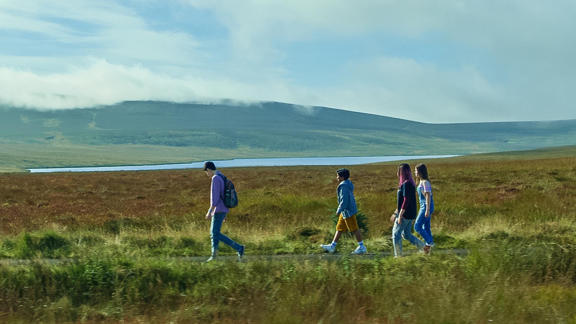 Blauer Himmel und eine weite nordirische Landschaft mit See und einer Hügelkette im Hintergrund: mittendurch laufen Louis, Glen, Kaz und Meg hintereinander auf einer kleinen Straße entlang.