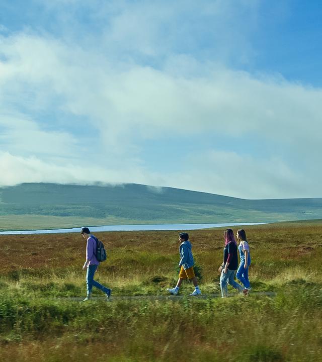 Blauer Himmel und eine weite nordirische Landschaft mit See und einer Hügelkette im Hintergrund: mittendurch laufen Louis, Glen, Kaz und Meg hintereinander auf einer kleinen Straße entlang.
