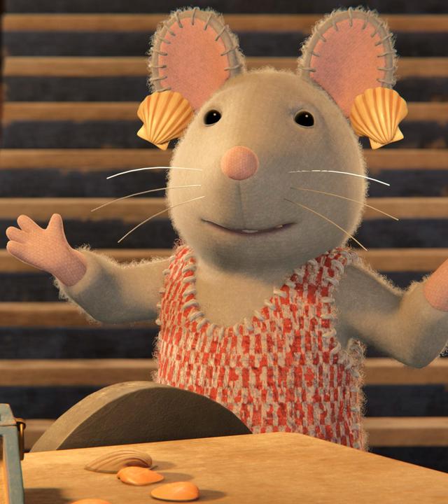 Julia steht mit ausgebreiteten Armen in einem Raum vor einem Tisch, auf dem eine kleine Holzkiste steht, in der Muscheln liegen. Neben der Kiste liegen ebenfalls ein paar Muscheln. Hinter Julia führt eine steile Treppe nach oben. Julia hat sich aus Spaß Muscheln als Ohrringe an die Ohren gehängt.