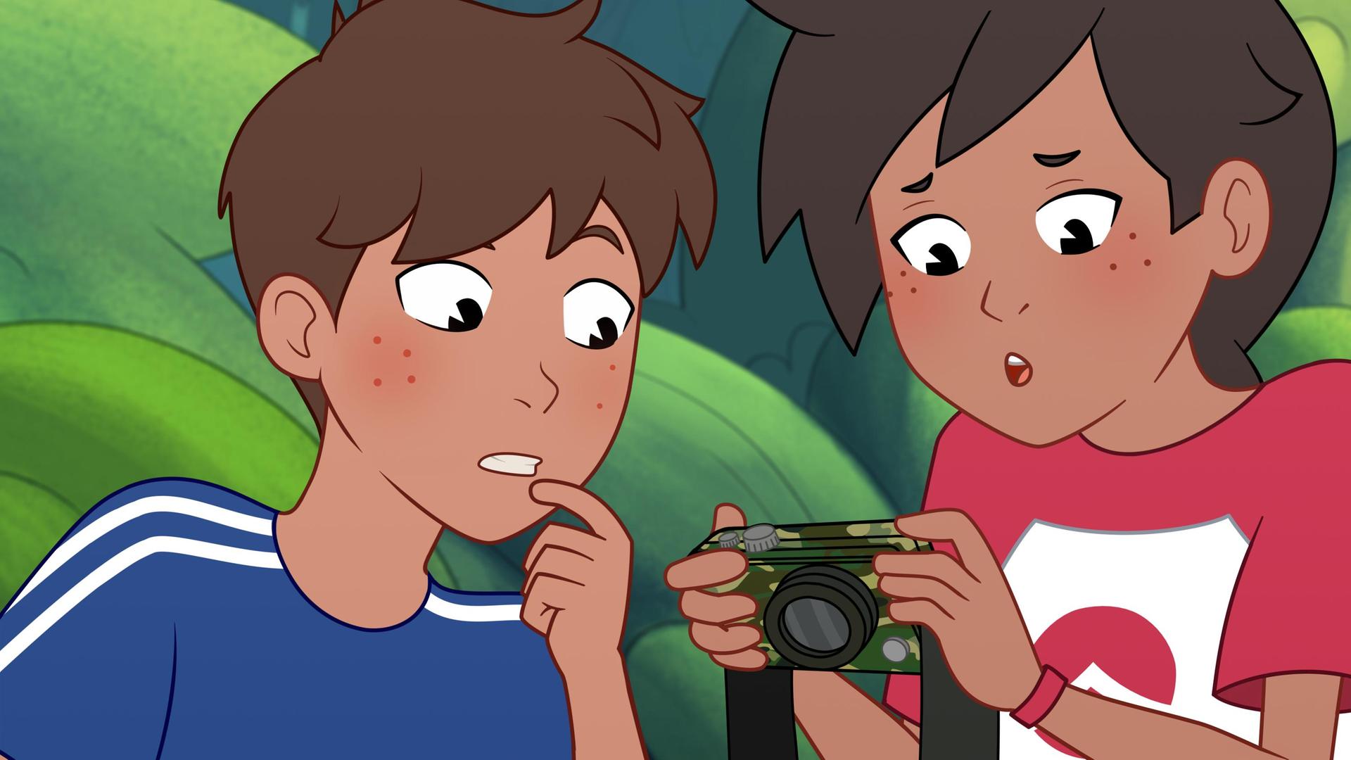 Josefine hält eine Kamera in grünen Tarnfarben in der Hand und schaut erschrocken darauf. Links neben ihr steht ihr Bruder Paul, der ebenfalls erschrocken darauf schaut. Er trägt ein blaues T-Shirt und Josefine ein rot-weißes T-Shirt. Im Hintergrund stehen Bäume.