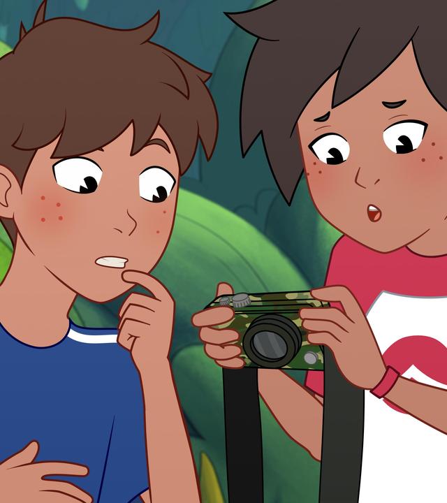 Josefine hält eine Kamera in grünen Tarnfarben in der Hand und schaut erschrocken darauf. Links neben ihr steht ihr Bruder Paul, der ebenfalls erschrocken darauf schaut. Er trägt ein blaues T-Shirt und Josefine ein rot-weißes T-Shirt. Im Hintergrund stehen Bäume.