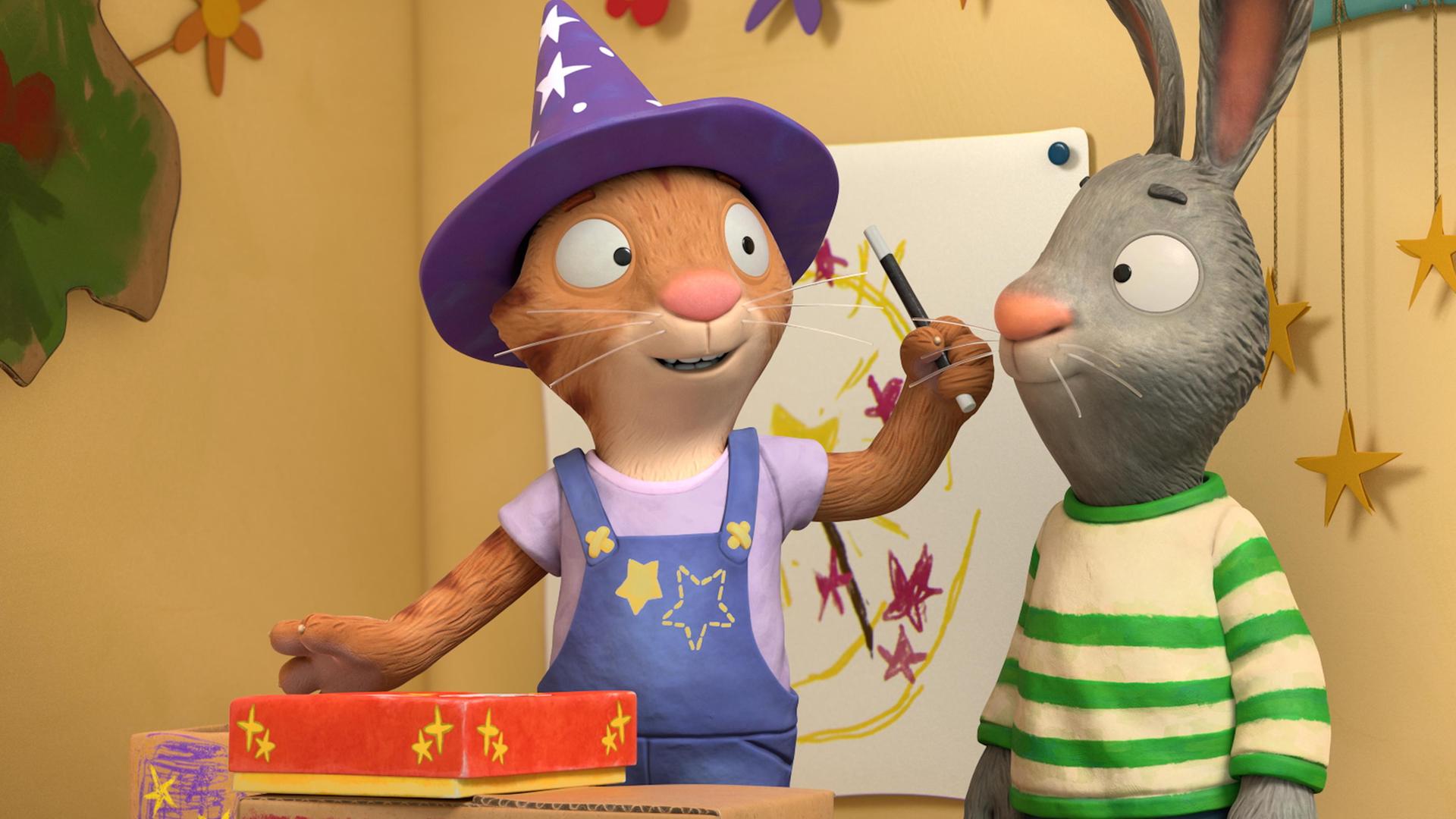 Frankie, die Katze gibt eine Zaubervorstellung für ihre Freunde. Sie trägt einen Zauberhut mit Sternen und Punkten, hält einen Zauberstab in ihrer Hand und ein geschlossenes Kästchen steht vor ihr auf der selbst gebastelten Bühne aus bunt angemalten Kartons.  Während sie ihren Zauberstab schwingt, dreht sie sich leicht zu Pip um, der direkt neben ihr steht.  Pip das Kaninchen blickt ihr neugierig zu.