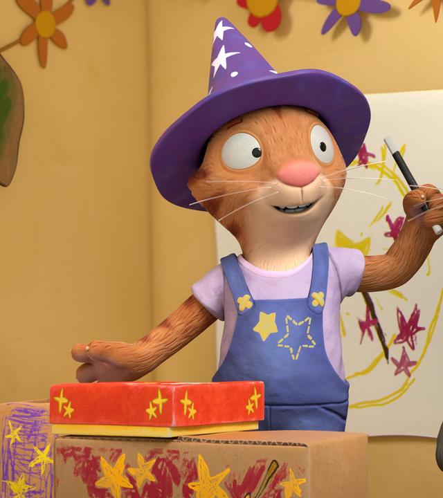 Frankie, die Katze gibt eine Zaubervorstellung für ihre Freunde. Sie trägt einen Zauberhut mit Sternen und Punkten, hält einen Zauberstab in ihrer Hand und ein geschlossenes Kästchen steht vor ihr auf der selbst gebastelten Bühne aus bunt angemalten Kartons.  Während sie ihren Zauberstab schwingt, dreht sie sich leicht zu Pip um, der direkt neben ihr steht.  Pip das Kaninchen blickt ihr neugierig zu.