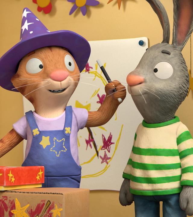 Frankie, die Katze gibt eine Zaubervorstellung für ihre Freunde. Sie trägt einen Zauberhut mit Sternen und Punkten, hält einen Zauberstab in ihrer Hand und ein geschlossenes Kästchen steht vor ihr auf der selbst gebastelten Bühne aus bunt angemalten Kartons.  Während sie ihren Zauberstab schwingt, dreht sie sich leicht zu Pip um, der direkt neben ihr steht.  Pip das Kaninchen blickt ihr neugierig zu.