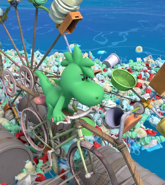 Grisu sitzt auf einem Fahrrad. Dieses wird von einem schaufelradartigen Mechanismus angerieben. Grisus Fahrrad schwimmt im Meer auf leeren Fässern und rings um ihn herum liegt ganz viel Müll im Wasser.