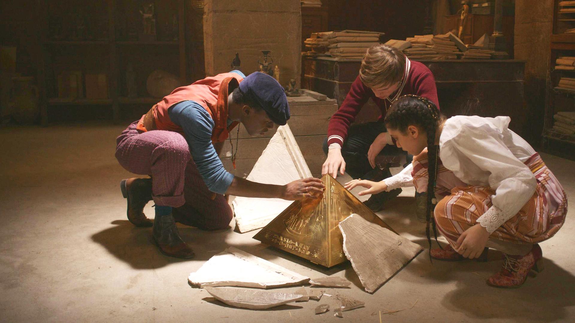 In einem dunklen Raum: Will, Henry und Safiya knien vor dem Pyramidion, von dem die äußere Hülle weggesplittert ist. Sie greifen alle drei mit ihrer rechten Hand vorsichtig darauf.