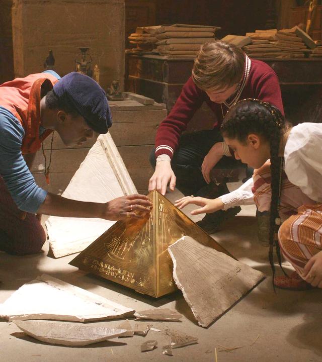 In einem dunklen Raum: Will, Henry und Safiya knien vor dem Pyramidion, von dem die äußere Hülle weggesplittert ist. Sie greifen alle drei mit ihrer rechten Hand vorsichtig darauf.