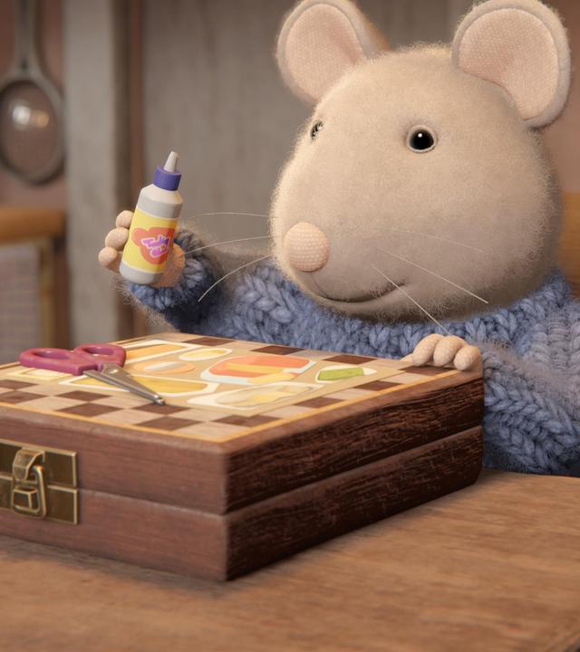 Sam sitzt in einer Wohnung auf einem Holzstuhl an einem Holztisch. Vor ihm auf dem Tisch liegt ein Schachbrettkasten. Darauf liegen ein Bild und eine Schere. Sam hält eine Flasche mit Flüssigkleber in der rechten erhobenen Hand und schaut auf das Bild vor sich.