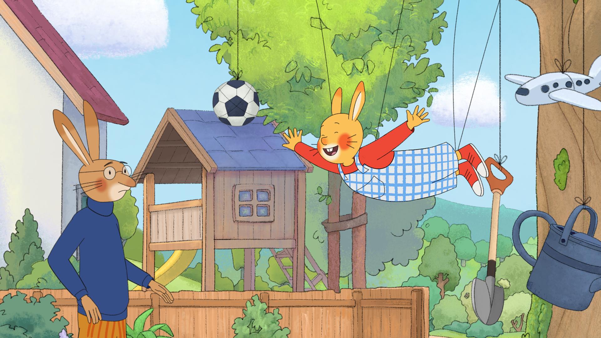 Karlchen hängt mit ausgebreiteten Armen an Schnüren, die an einem Baum befestigt sind. An Schnüren hängen auch ein Fußball, ein Spaten, eine Gießkanne und ein Spielzeugflugzeug. Karlchens Papa schaut skeptisch auf seinen an Sohn. 