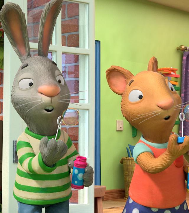 Die beiden allerbesten Freunde Pip das Kaninchen und Posy die Maus stehen draußen vor der geöffneten Terassentür und machen einen Seifenblasen-Wettbewerb. Sie stehen nebeneinander, halten jeweils ein Fläschchen mit Seifenblasenflüssigkeit in ihrer Hand und pusten vorsichtig. Die Seifenblasen der beiden sind fast gleich groß.