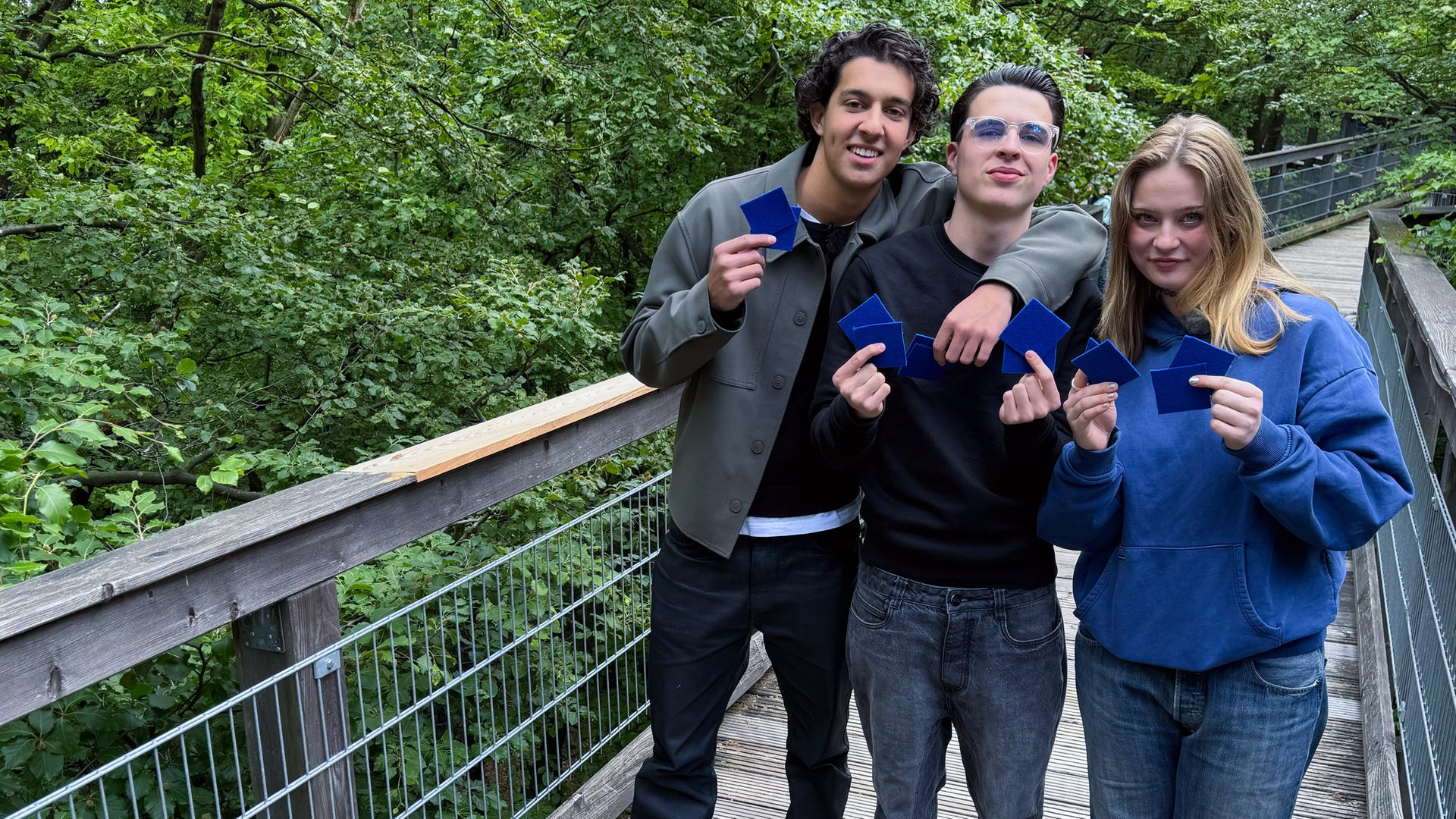 Drei Jugendliche stehen auf einer Art Holzbrücke, umgeben von Bäumen und Blättern. Es handelt sich um einen Baumwipfelpfad. Die drei halten blaue Karten in die Kamera und sehen entschlossen aus, die Challenge zu gewinnen.