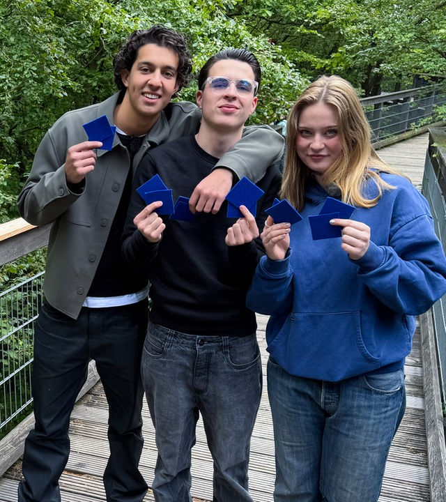 Drei Jugendliche stehen auf einer Art Holzbrücke, umgeben von Bäumen und Blättern. Es handelt sich um einen Baumwipfelpfad. Die drei halten blaue Karten in die Kamera und sehen entschlossen aus, die Challenge zu gewinnen.
