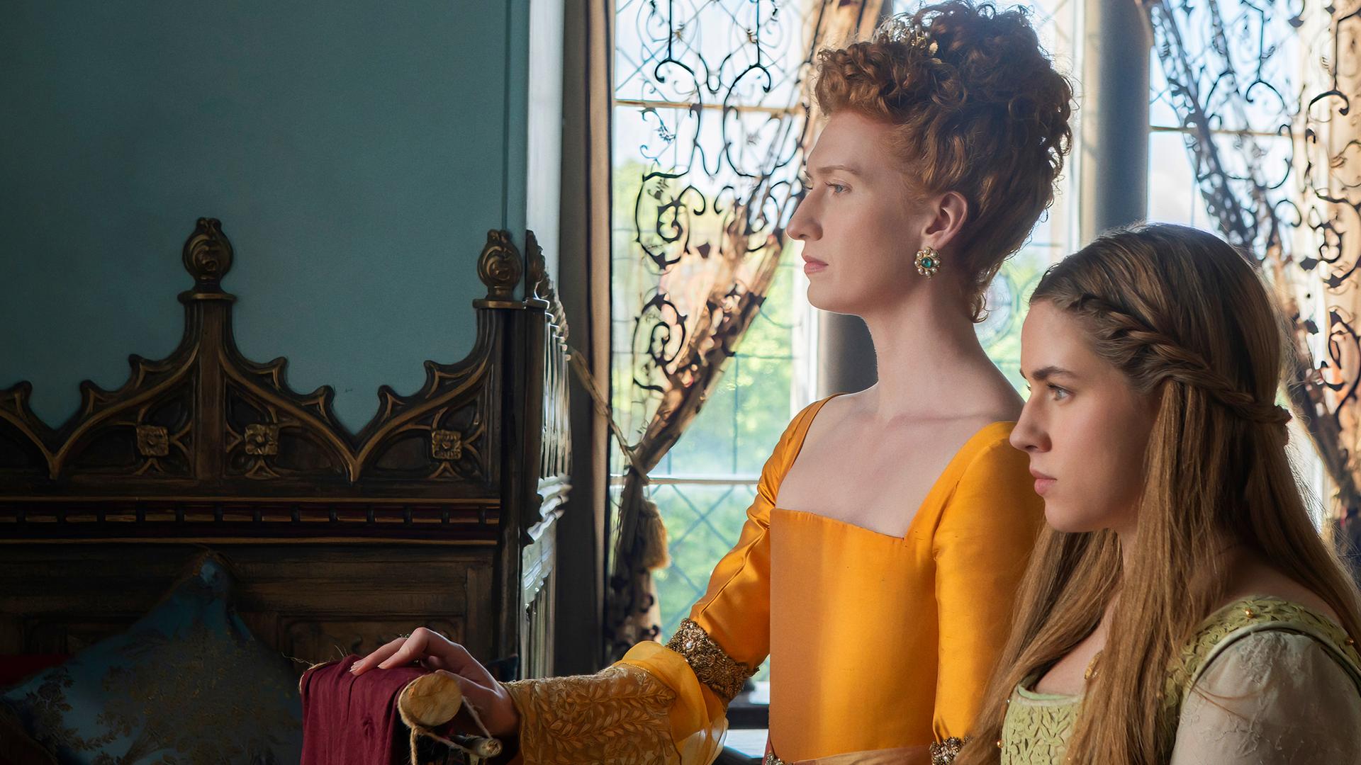 In einem Saal im Schloss stehen die Königin und Grace nebeneinander und blicken aus dem Fenster. Die Königin trägt ein gelb-oranges Kleid und hat die Haare kunstvoll hochgesteckt.
