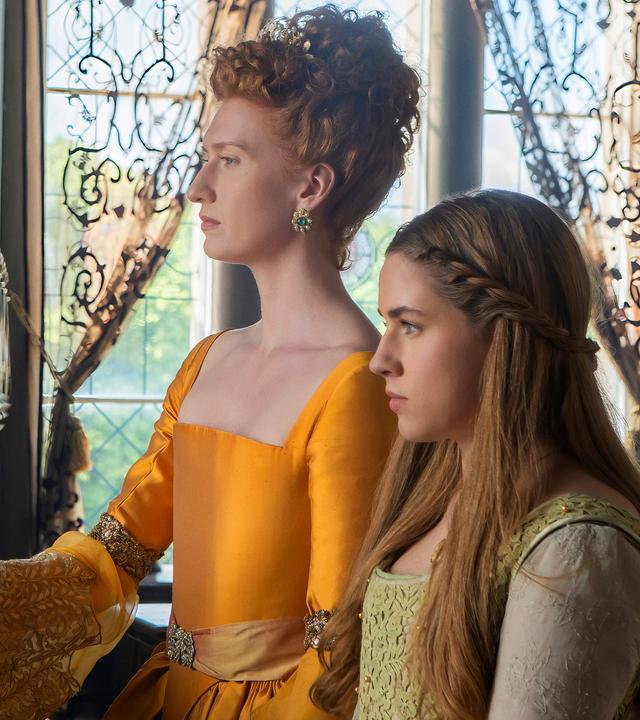 In einem Saal im Schloss stehen die Königin und Grace nebeneinander und blicken aus dem Fenster. Die Königin trägt ein gelb-oranges Kleid und hat die Haare kunstvoll hochgesteckt.