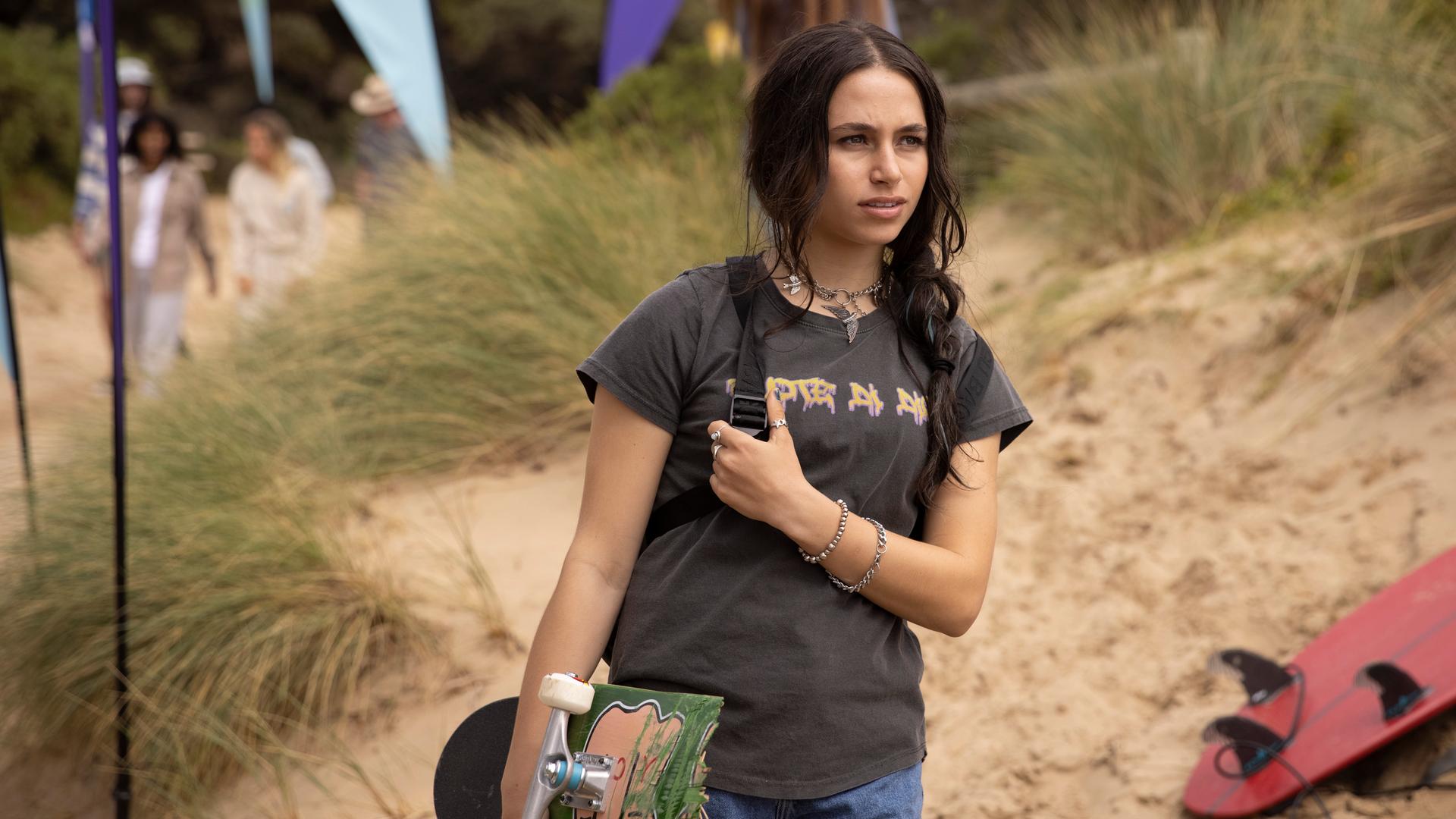 Am Strand: Summer steht mit ihrem Skateboard unter dem rechten Arm am Strand. Sie blickt skeptisch nach vorne. Neben ihr im Sand liegen Surfbretter und im Hintergrund sind Sand, Gräser und Menschen zu sehen.