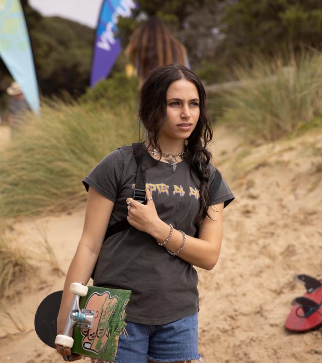Am Strand: Summer steht mit ihrem Skateboard unter dem rechten Arm am Strand. Sie blickt skeptisch nach vorne. Neben ihr im Sand liegen Surfbretter und im Hintergrund sind Sand, Gräser und Menschen zu sehen.