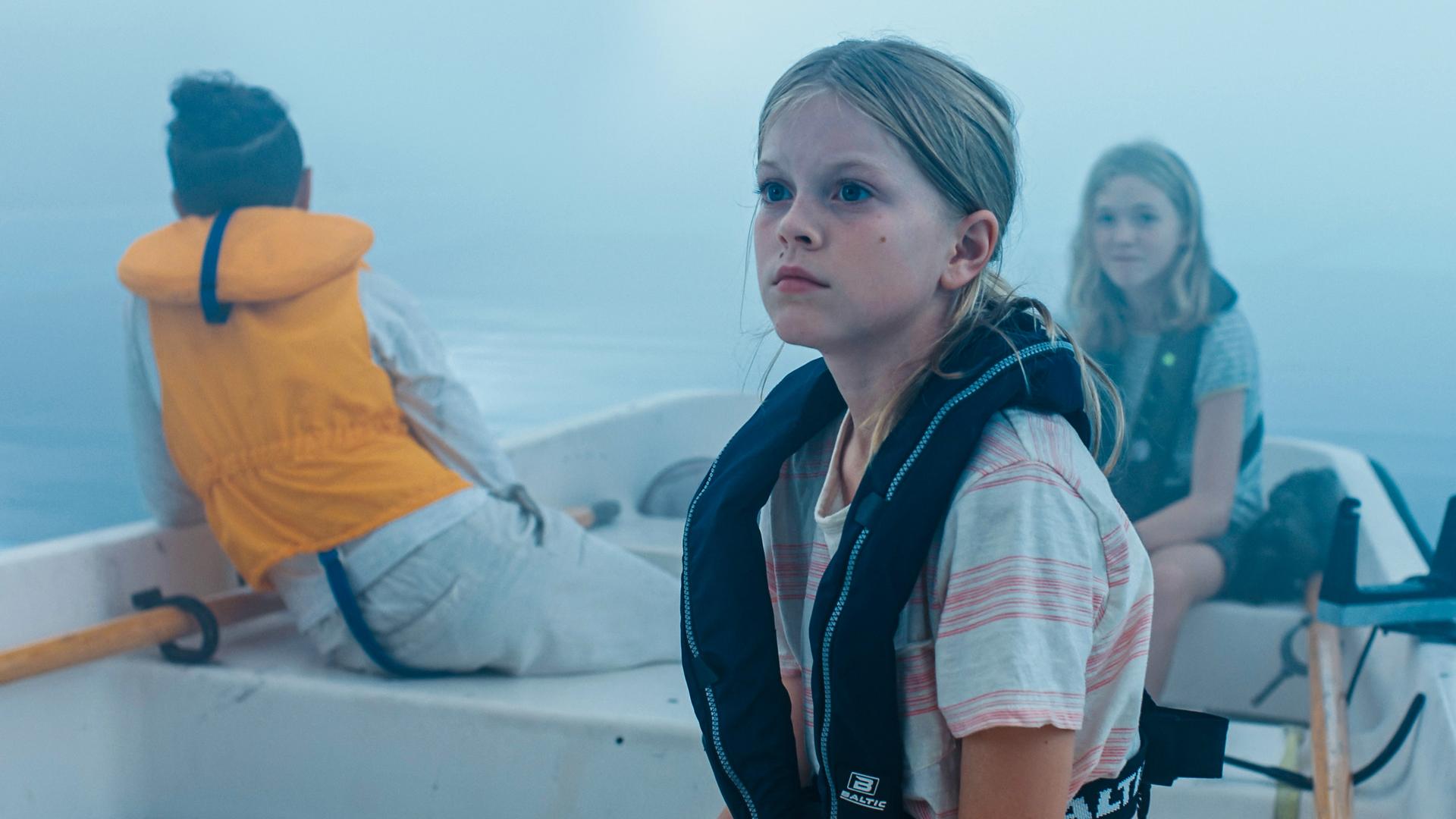 Elliot, Elin und Julia sitzen im dichten Nebel auf einem Gewässer in einem kleinen Ruderboot. Im Vordergrund sitzt Elin mit einer dunklen Schwimmweste und blickt zur Seite. Hinter ihr sitzt sitzt Elliot, die eine orangefarbene Schwimmweste trägt und nur von hinten zu sehen ist und in den Nebel schaut. Auf der anderen Seite sitzt sitzt Julia, die ebenfalls nach vorne schaut.
