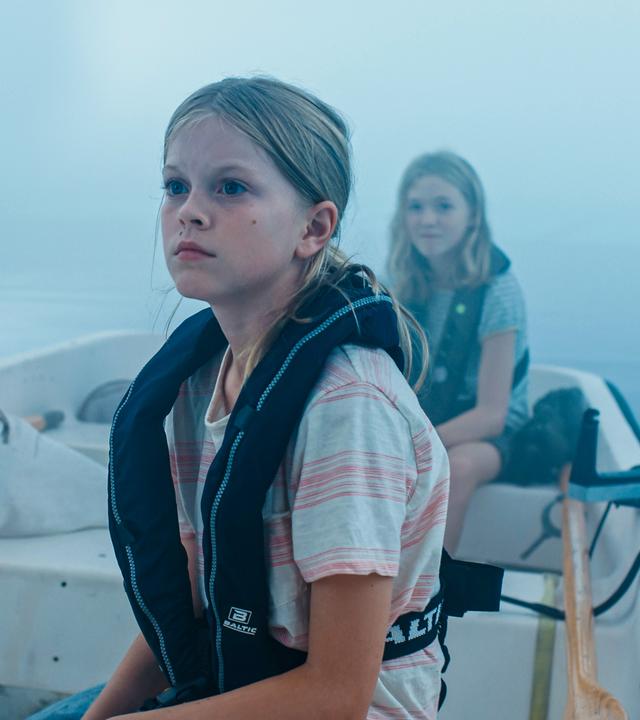 Elliot, Elin und Julia sitzen im dichten Nebel auf einem Gewässer in einem kleinen Ruderboot. Im Vordergrund sitzt Elin mit einer dunklen Schwimmweste und blickt zur Seite. Hinter ihr sitzt sitzt Elliot, die eine orangefarbene Schwimmweste trägt und nur von hinten zu sehen ist und in den Nebel schaut. Auf der anderen Seite sitzt sitzt Julia, die ebenfalls nach vorne schaut.