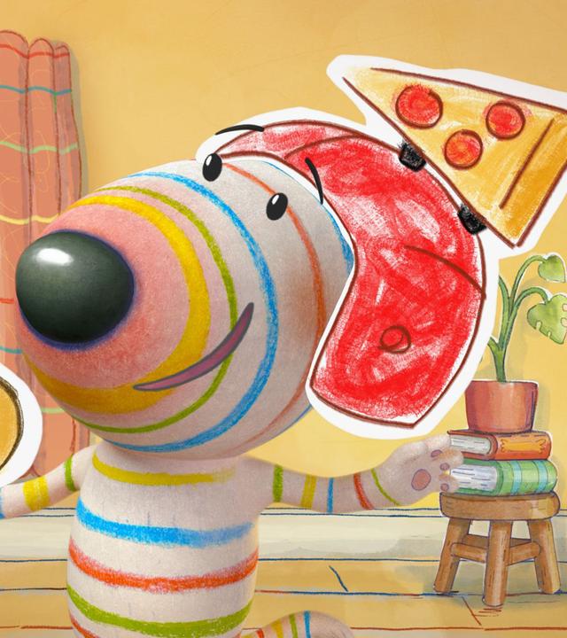 Dylan steht freudestrahlend in der Mitte seines Wohnzimmers. Auf dem Kopf trägt er einen Helm, auf dessen Spitze ein Stück Pizza befestigt ist. In einer Pfote balanciert er eine Salamipizza.