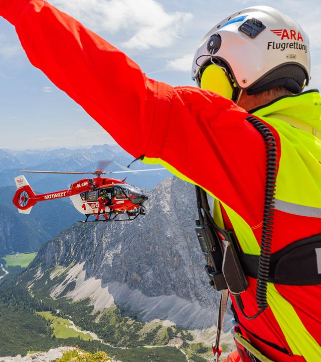 Alpenpanorama: Im Vordergrund hebt ein Mann die Arme seitlich wie zu einem „Y“, um den im Hintergrund anfliegenden Hubschrauber zur sicheren Landung zu weisen.