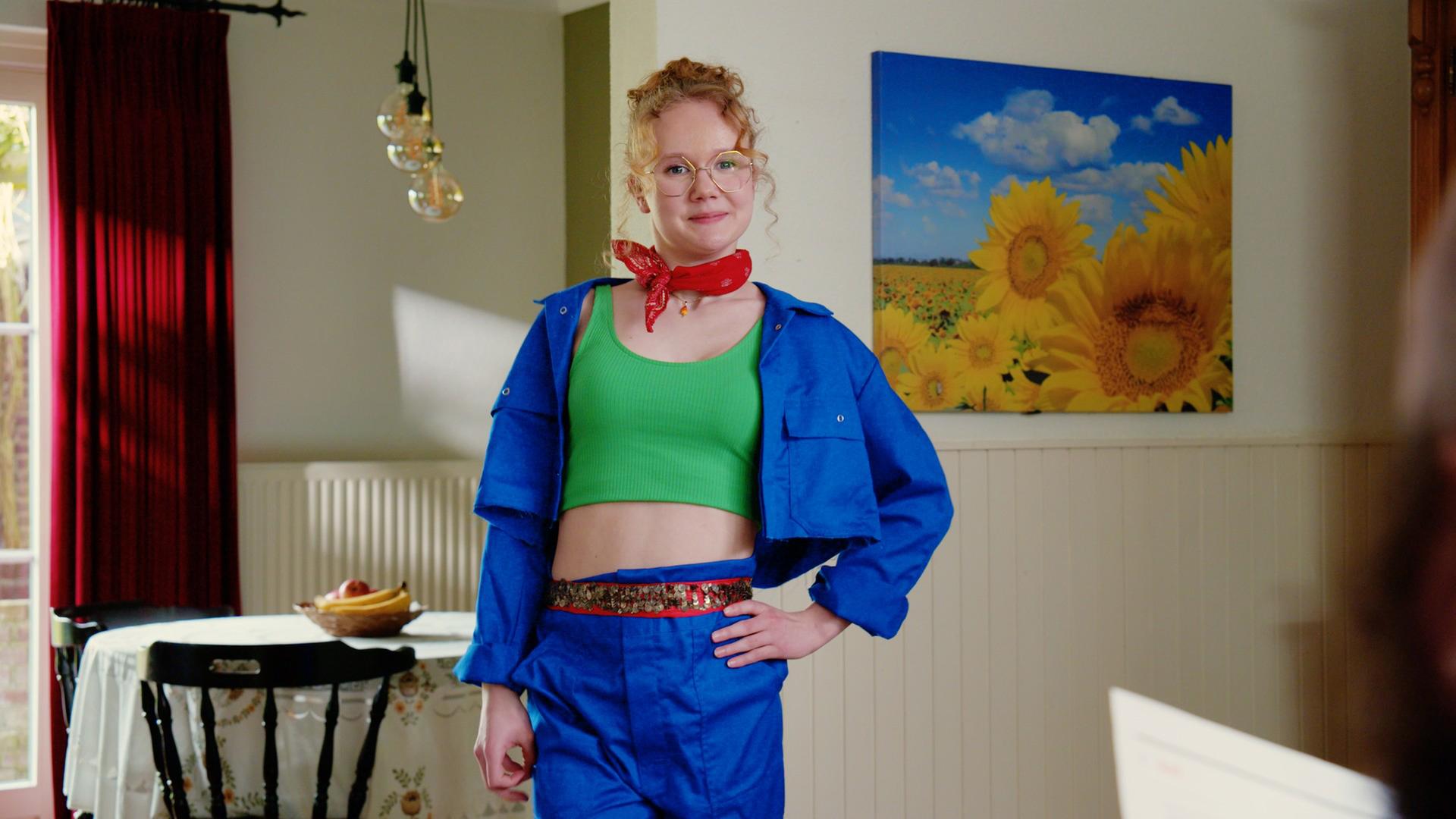 Lena steht mit blauer Hose und Jacke sowie einem grünen bauchfreien Top in einem Wohn-Esszimmer. 