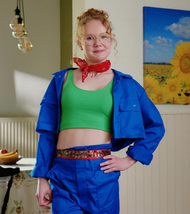 Lena steht mit blauer Hose und Jacke sowie einem grünen bauchfreien Top in einem Wohn-Esszimmer. 