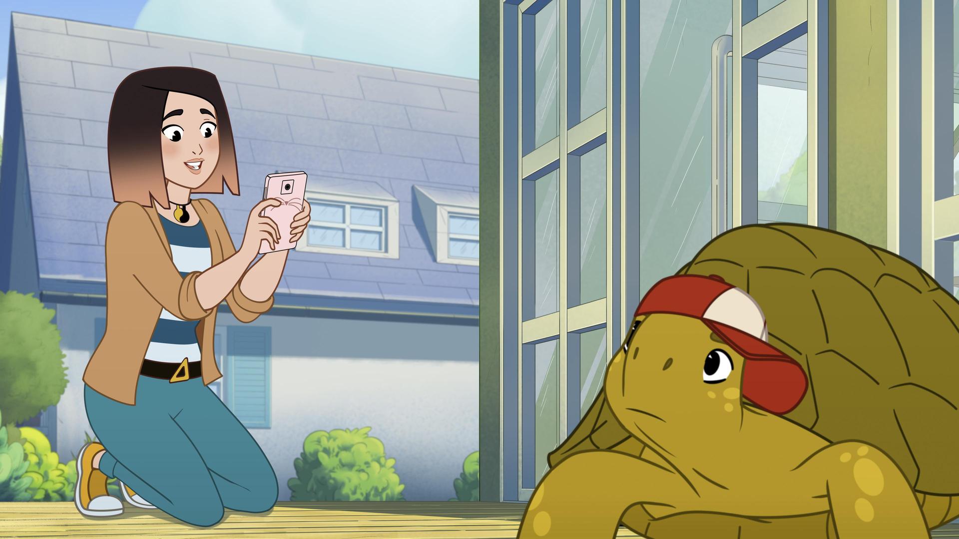 Die grüne Schildkröte Törtel trägt mit verunsichertem Blick eine rot-weiße Kappe auf dem Kopf, während weiter hinten Alina ein Foto mit ihrem rosafarbenen Handy von ihm macht. Alina ist eine junge Frau mit kurzen braun-blonden Haaren. Im Hintergrund steht ein Wohnhaus umgeben von grünen Büschen.