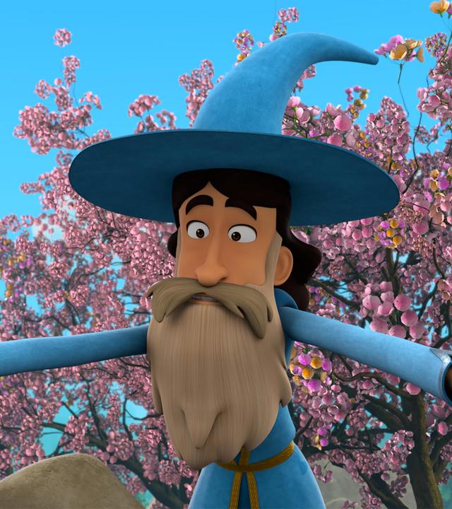 Ein Mann mit dunklem Haar und hellem, langen Bart und Schnurrbart in der blau-goldfarbenen Kleidung von Merlin und dessen Zauberhut, beugt sich mit weit ausgebreiteten Armen zu etwas hinab. Hinter ihm sind Obstbäume, die in voller, rosafarbener Blüte stehen.