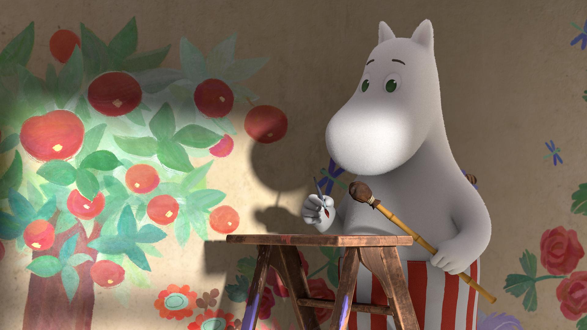 Muminmama steht auf einer Leiter und hält einen Malerpinsel in ihrer Hand. Im Hintergrund ist das von ihr an die Wand gemalte Wandbild zu erkennen: ein Apfelbaum und eine Blumenwiese.