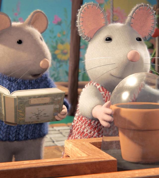 Sam und Julia stehen vor einem Regal auf der Terrasse. Dort steht ein Blumentopf, über den Julia eine Glasglocke stülpt. Sam steht neben ihr und hält ein geöffnetes Buch in der Hand. Er erzählt Julia etwas.