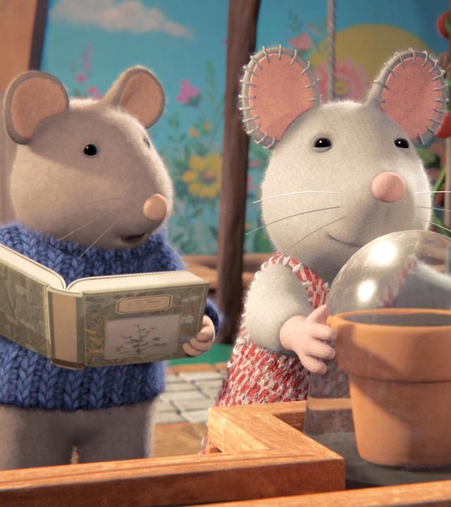 Sam und Julia stehen vor einem Regal auf der Terrasse. Dort steht ein Blumentopf, über den Julia eine Glasglocke stülpt. Sam steht neben ihr und hält ein geöffnetes Buch in der Hand. Er erzählt Julia etwas.