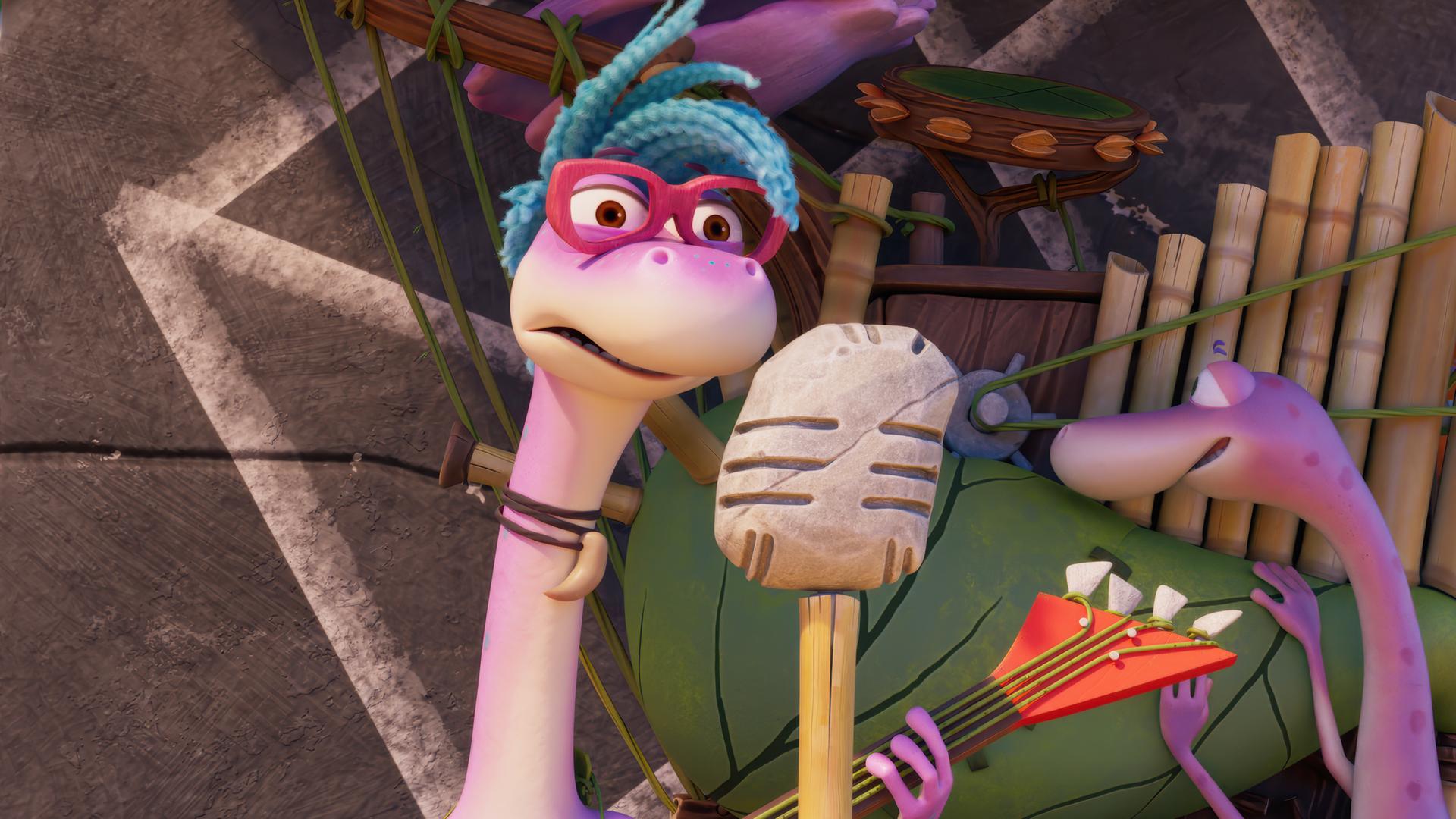 Das Dinosauriermädchen Magma mit der coolen Brille und den blauen Haaren spielt mit einer roten E-Gitarre auf einer Bühne. Vor ihr steht ein steinzeitliches Mikrofon auf einem Stativ. Im Hintergrund steht Minus Drei vor einer merkwürdigen Maschine, die aussieht wie ein Blasebalg mit Orgelpfeifen.