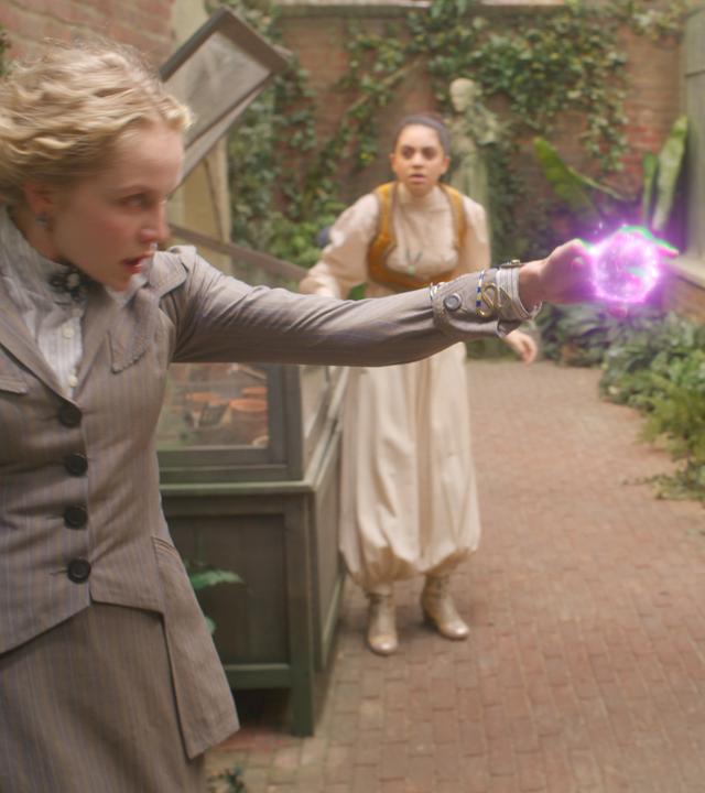 Nigella steht Theodosia gegenüber. Sie hält ihren linken Arm nach oben und richtet ihre Hand, aus der ein leuchtender Strahl kommt, gegen Theodosia. Ihr Blick ist böse. Theodosia hält mit erschrockenem Blick ihre Hände leicht nach oben Richtung Strahl. Im Hintergrund sind Safiya und Henry zu sehen, die das Ganze mit geschocktem Blick beobachten.