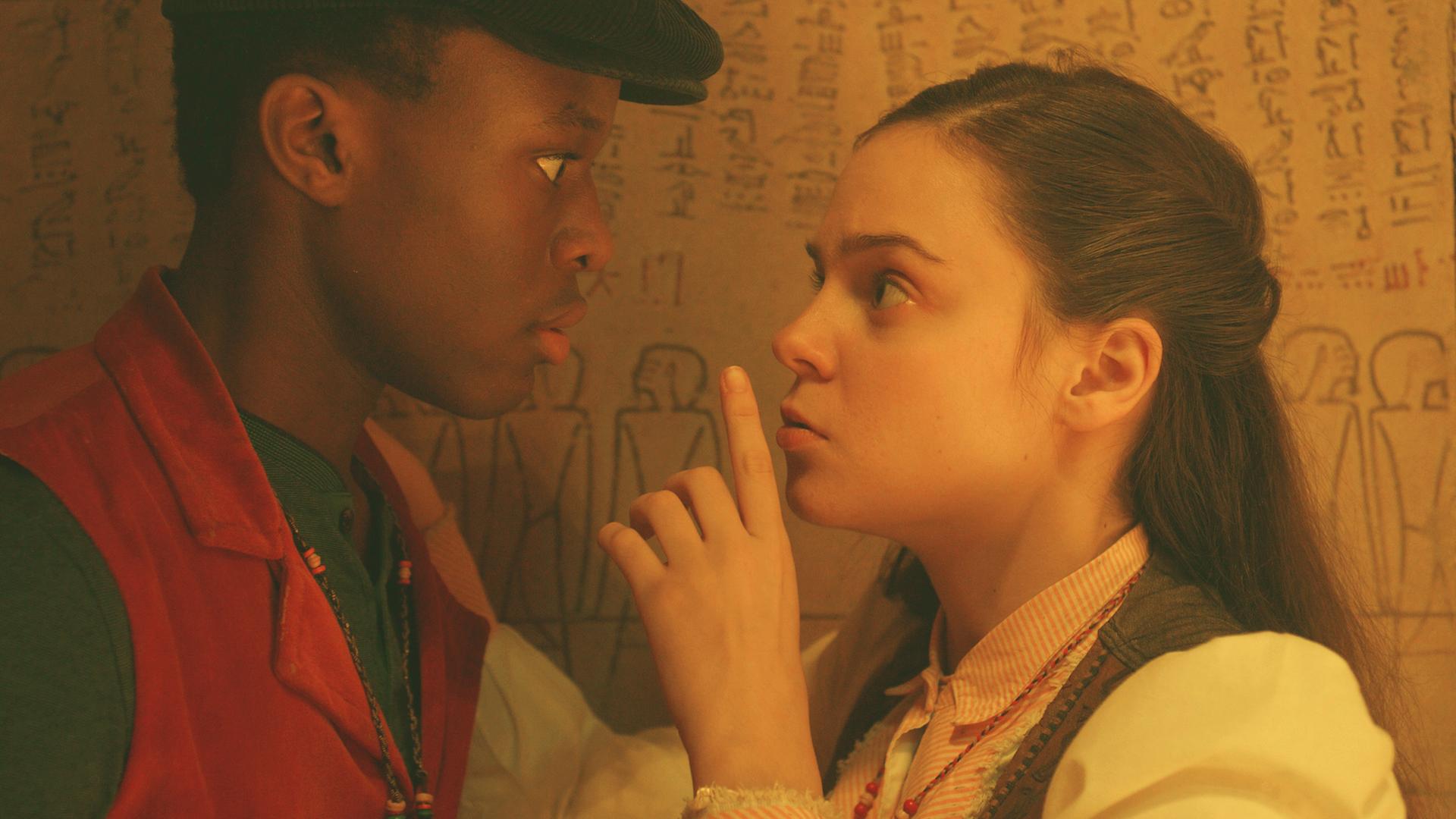 Will und Theodosia stehen sich gegenüber vor einer Wand mit Hieroglyphen und sehen sich in die Augen. Theodosia hält sich den Zeigefinger der linken Hand zum Zeichen für "Still sein!" vor den Mund.