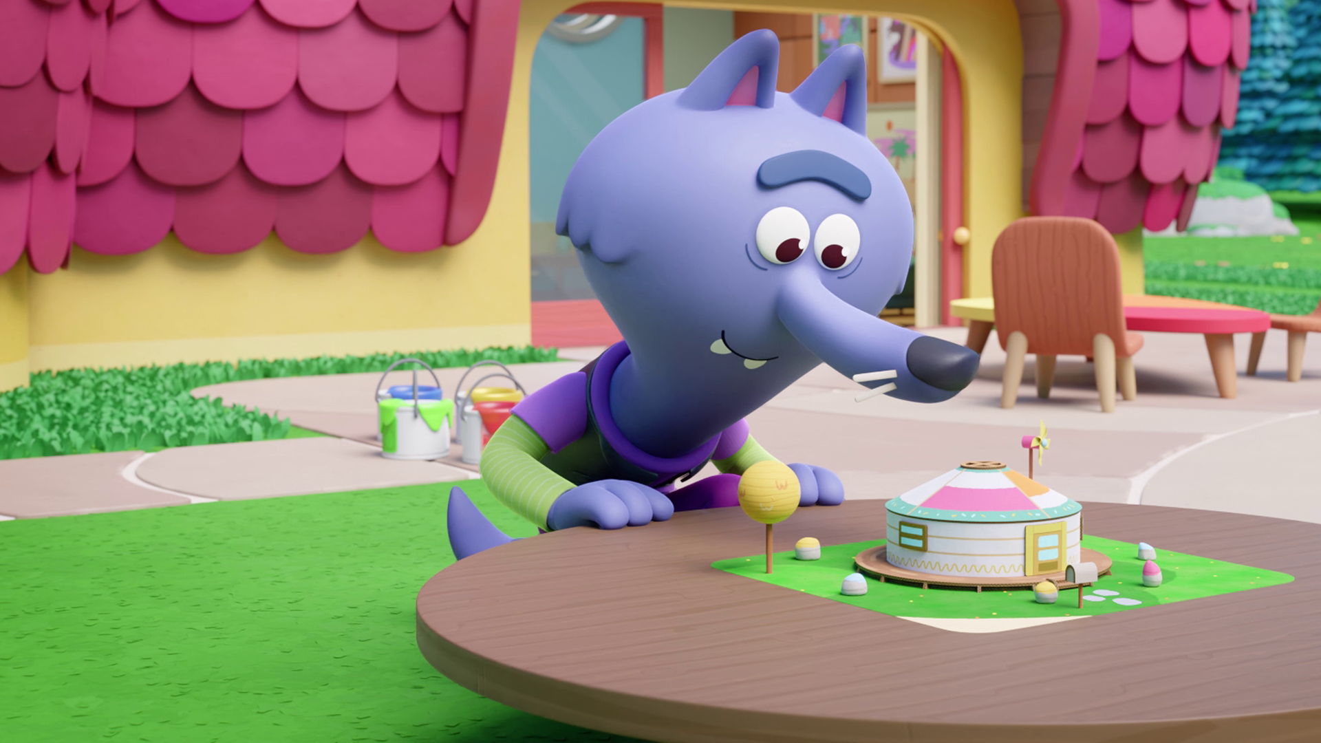 Wolf Wilf steht an einem Tisch, auf dem ein rundes Miniatur-Zelt detailgetreu aufgebaut ist. Fasziniert betrachtet er es. Im Hintergrund sehen wir ein Haus mit pinken Dachziegeln und eine kleine Terrasse davor.