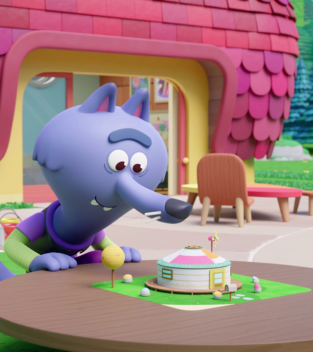 Wolf Wilf steht an einem Tisch, auf dem ein rundes Miniatur-Zelt detailgetreu aufgebaut ist. Fasziniert betrachtet er es. Im Hintergrund sehen wir ein Haus mit pinken Dachziegeln und eine kleine Terrasse davor.