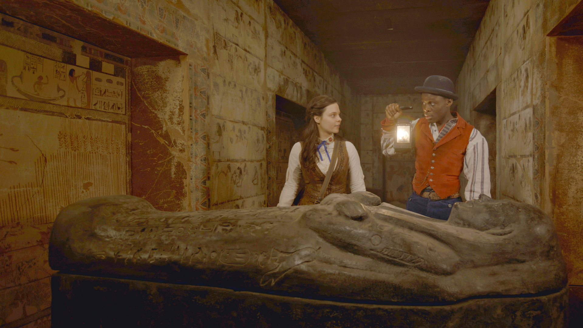 Theodosia und Will erkunden einen unterirdischen Gang. Will hält eine Petroleumlampe in der rechten Hand hoch und beleuchtet damit einen Sarkophag.
