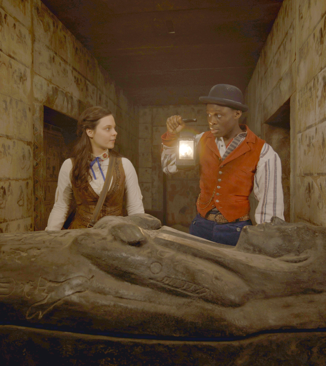 Theodosia und Will erkunden einen unterirdischen Gang. Will hält eine Petroleumlampe in der rechten Hand hoch und beleuchtet damit einen Sarkophag.