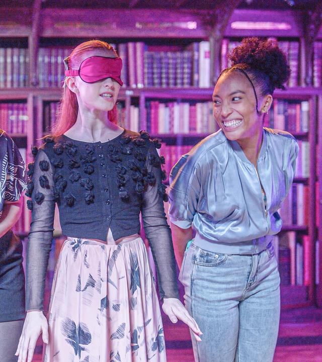 In einem nur spärlich beleuchteten Raum an dessen Rückwand Regale voller Bücher stehen: Die vier Freundinnen stehen in einer Reihe nebeneinander. Die aufgeregte Mia hat die Augen mit einer Schlafmaske verbunden. Lola und Amy verkneifen sich ein Kichern und Cece strahlt mit breitem Grinsen Amy an.