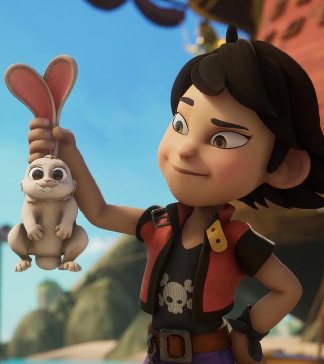 Draußen am Strand steht Zoe. Sie hat ihren linken Arm in die Hüfte gestemmt und in der rechten Hand hält sie ein niedliches Kaninchen an beiden Ohren hoch und schaut es freundlich an.