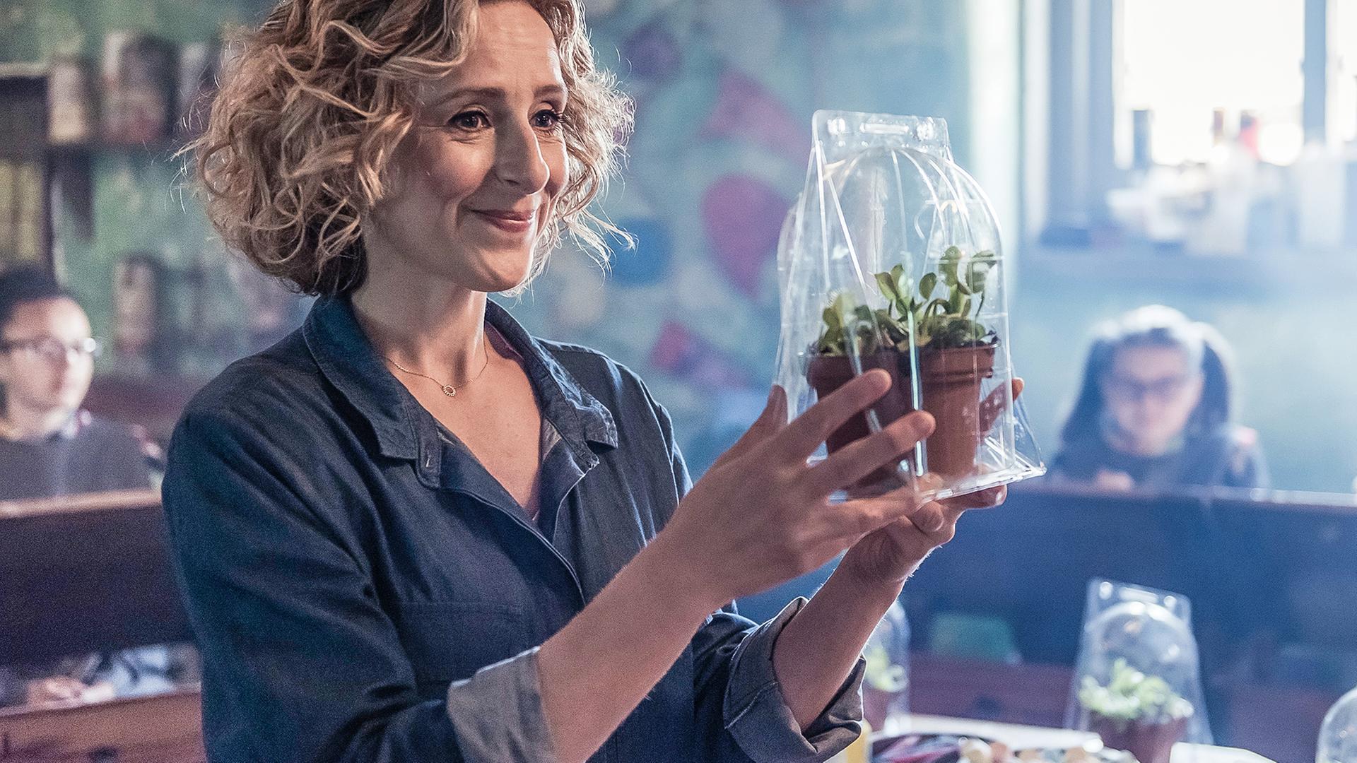 Julie, die neue Kunstlehrerin, steht in einem Klassenraum und schaut lächelnd eine Pflanze an, die sie in den Händen hochhält.