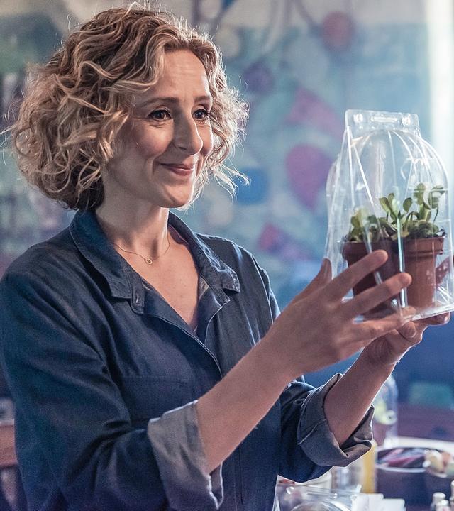 Julie, die neue Kunstlehrerin, steht in einem Klassenraum und schaut lächelnd eine Pflanze an, die sie in den Händen hochhält.